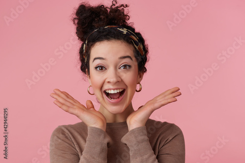 Portrait of attractive, joyful girl with dark curly hair bun. Wearing headband, earrings and brown sweater. Has make up. Keeps palms spread. Watching at the camera isolated over pastel pink background