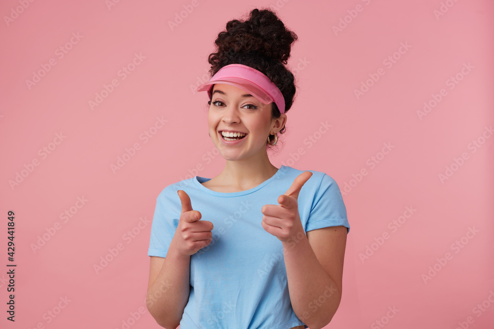 Flirty woman, beautiful girl with dark curly hair bun. Wearing pink visor, earrings and blue t-shirt. Has make up. Pointing fingers at you. Watching at the camera isolated over pastel pink background