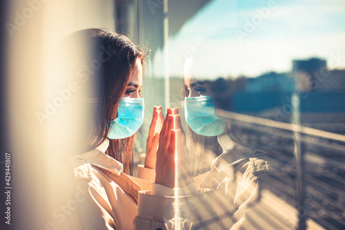 Girl with medical mask is looking out of the window during pandemic of Coronavirus Covid-19