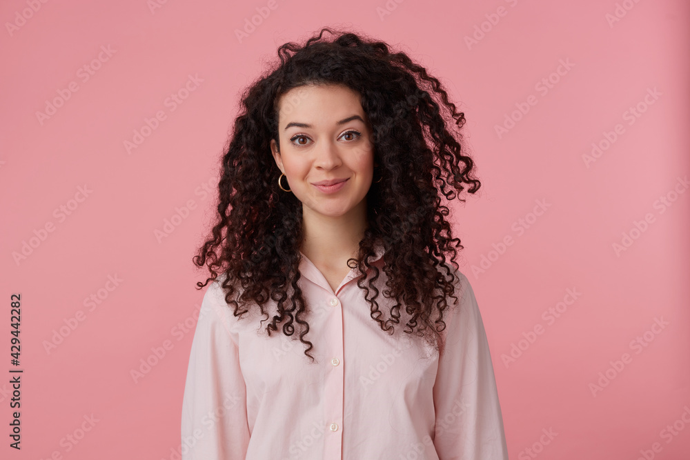 Charming girl, happy looking woman with dark long hair. Wearing earrings and pastel pink shirt. Has make up. People and emotion concept. Watching at the camera isolated over pastel pink background Charming girl, happy looking woman with dark long hair. Wearing earrings and pastel pink shirt. Has make up. People and emotion concept. Watching at the camera isolated over pastel pink background