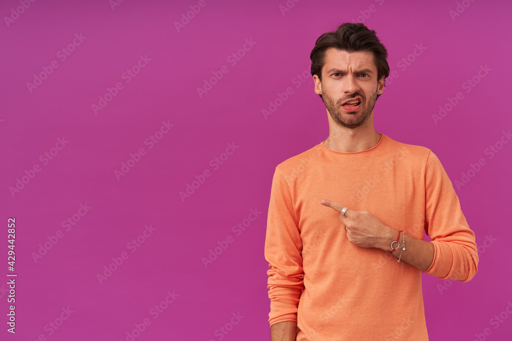 Portrait of frowning, displeased male with brunette hair and bristle. Wearing orange sweater. Watching at the camera and pointing finger to the left at copy space, isolated over purple background Portrait of frowning, displeased male with brunette hair and bristle. Wearing orange sweater. Watching at the camera and pointing finger to the left at copy space, isolated over purple background