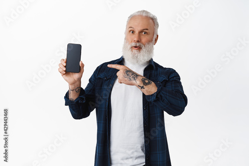 Impressed old tattooed guy pointing at smartphone screen, showing amazing new mobile app, display an application, standing over white background