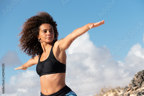 Multiracial young woman practicing yoga outdoors on open arms warrior pose II