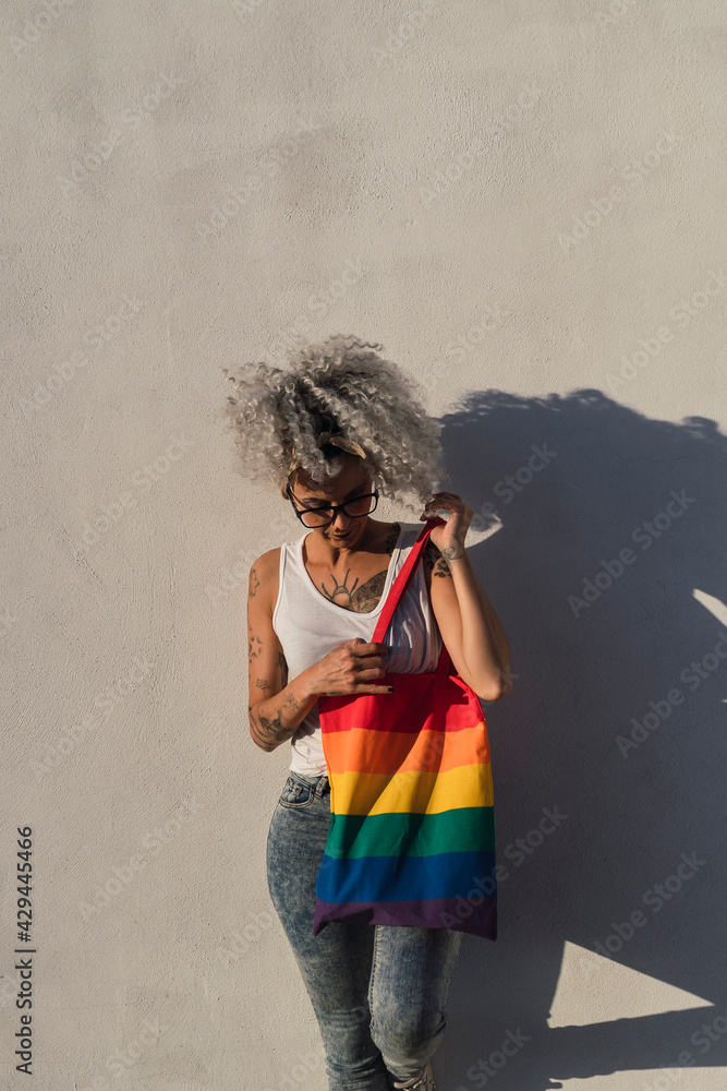 Chica delgada con pelo muy rizado delante de pared blanca posando feliz ocn una bolsa con la bandera de arco iris Chica delgada con pelo muy rizado delante de pared blanca posando feliz ocn una bolsa con la bandera de arco iris