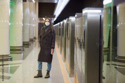 A woman in a medical face mask is waiting for a train and holding a smartphone.