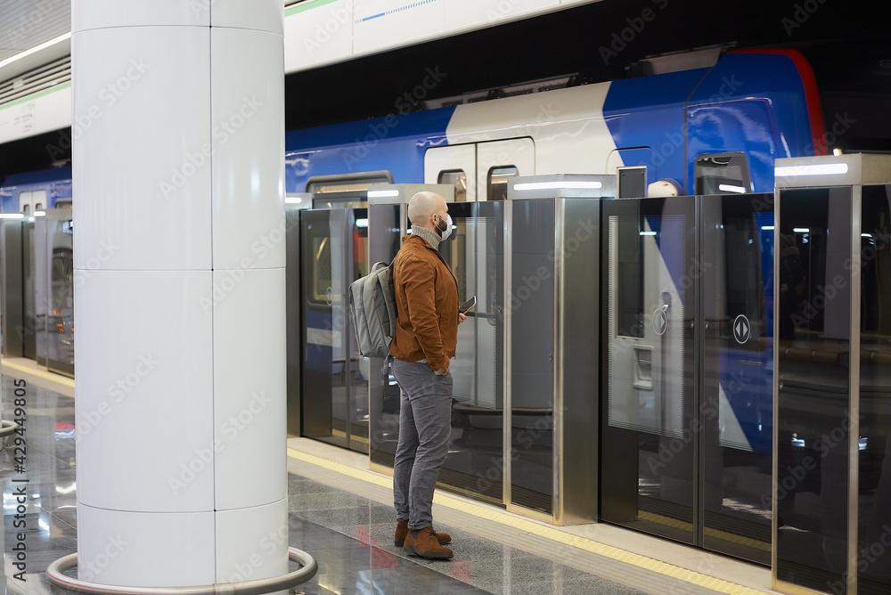 A man in a face mask is holding a smartphone while waiting for a subway train. A man in a face mask is holding a smartphone while waiting for a subway train.