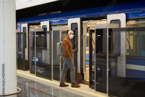 A man in a medical face mask is holding a smartphone while entering a subway car