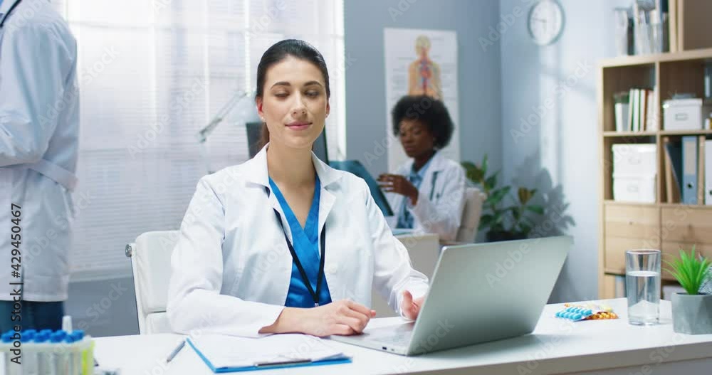 Portrait of happy joyful Caucasian young beautiful professional doctor woman working in cabinet typing on laptop sitting at desk in hospital looking at camera and smiling. Physician concept Portrait of happy joyful Caucasian young beautiful professional doctor woman working in cabinet typing on laptop sitting at desk in hospital looking at camera and smiling. Physician concept