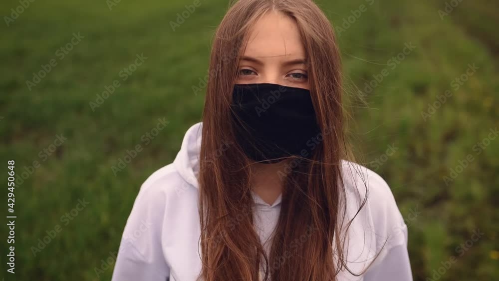 A girl in a white sweater and black mask puts on a hood and looks at the camera on a background of green grass. hair fluttering in the wind. pandemic covid-19. Coronavirus A girl in a white sweater and black mask puts on a hood and looks at the camera on a background of green grass. hair fluttering in the wind. pandemic covid-19. Coronavirus