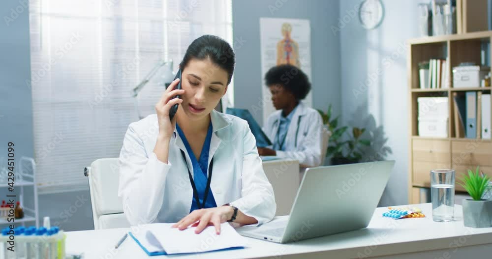 Portrait of Caucasian young beautiful professional doctor woman working in cabinet at desk in hospital speaking on smartphone on call with patient. Physician concept. healthcare medicine Portrait of Caucasian young beautiful professional doctor woman working in cabinet at desk in hospital speaking on smartphone on call with patient. Physician concept. healthcare medicine
