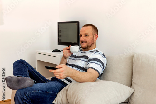 Cropped shot of a young man relaxing on the sofa, drinking coffee while watching tv at home during the day. Man watching tv while sitting on sofa in his living room. People and leisure concept.