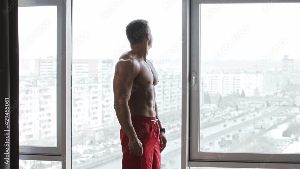 Muscular shirtless man standing on the background of panoramic windows and looking to the sides
