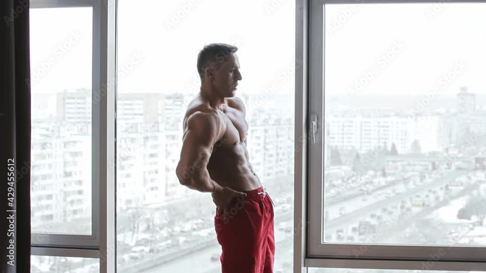 Muscular shirtless man standing on the background of panoramic windows and warming up his waist by the circular motions before the training Muscular shirtless man standing on the background of panoramic windows and warming up his waist by the circular motions before the training