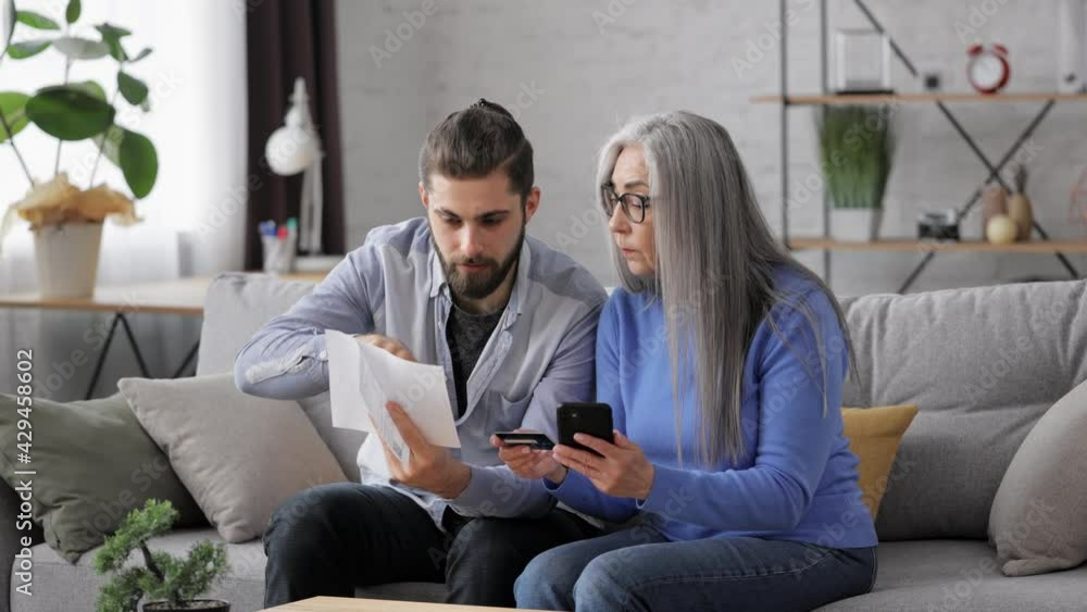 Adult son teaching his senior mother shopping online using smartphone. Young man helping aged mother paying bills online with credit card, making purchase in online store. Instant easy payments. Adult son teaching his senior mother shopping online using smartphone. Young man helping aged mother paying bills online with credit card, making purchase in online store. Instant easy payments.