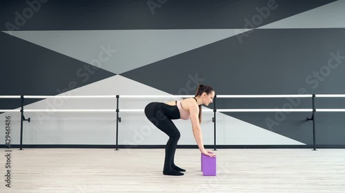 Side motion of beautiful fit caucasian woman wearing black sportswear stretching legs using small purple blocks on floor. Female dancer practicing in emapty hall. Stretching, gymnastics concept. photo