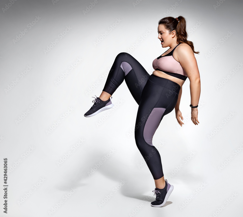 Girl practicing exercise, performs kick up. Photo of model with curvy  figure in fashionable sportswear on grey background. Sports motivation and  healthy lifestyle Stock Photo