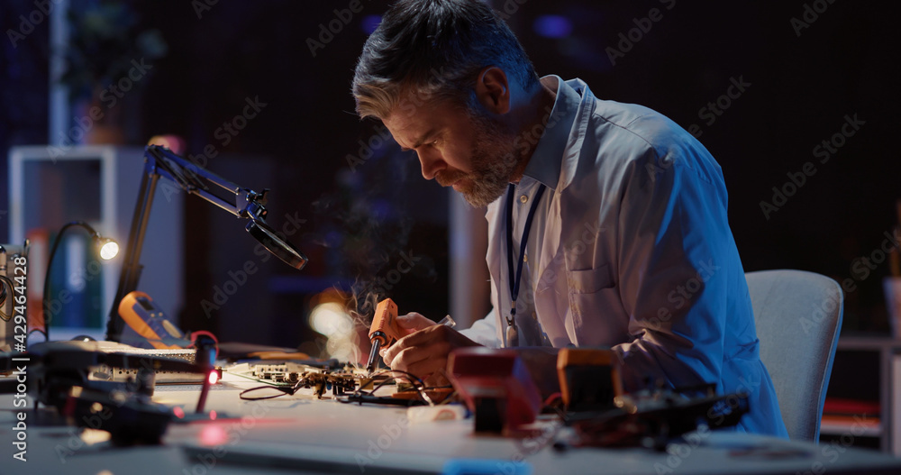 Caucasian adult engineer technician in lab coat sitting inside office soldering circuit board of broken drone using modern equipment working at night. Caucasian adult engineer technician in lab coat sitting inside office soldering circuit board of broken drone using modern equipment working at night.