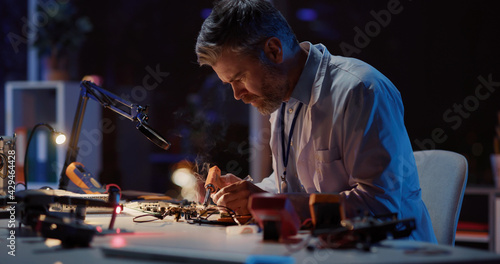 Caucasian adult engineer technician in lab coat sitting inside office soldering circuit board of broken drone using modern equipment working at night.