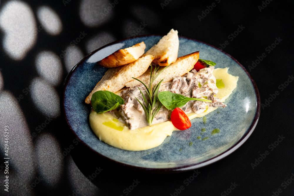 Meat in a creamy sauce with mashed potatoes and bread toast, painted with spinach leaves and cherry tomatoes, on a gray plate on a black background. Meat in a creamy sauce with mashed potatoes and bread toast, painted with spinach leaves and cherry tomatoes, on a gray plate on a black background.