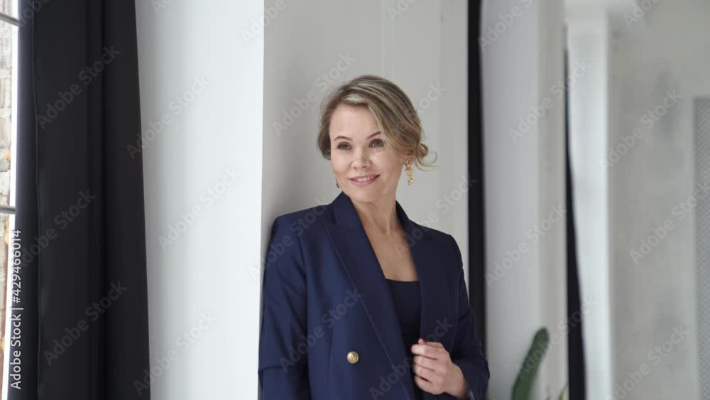 A blonde woman in a blue suit poses standing by a window in a photo studio. A blonde woman in a blue suit poses standing by a window in a photo studio.