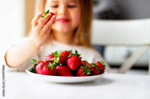 Cute little girl eating fresh strawberry in the kitchen. Healthy vitamin snack for kids. Ripe fresh berries. Harvest season. Natural vitamins .