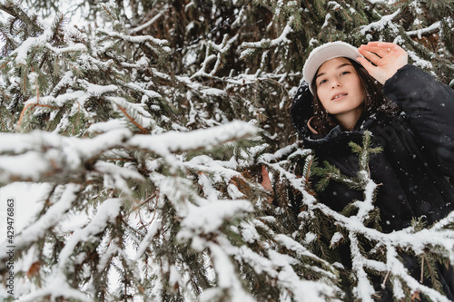 Photo of a girl standing on a background of a coniferous tree in the woods and closed by snow-covered branches of a Christmas tree