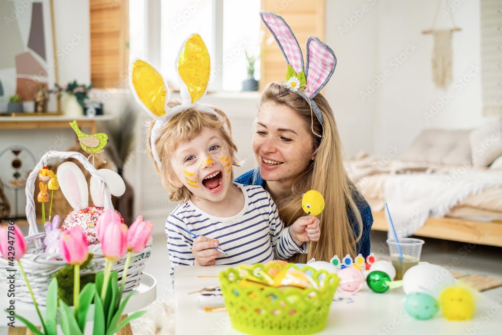 Beautiful happy son and mother in headbands with bunny ears decorate eggs with a brush and bright paints, prepare to celebrate Easter, have fun at home Beautiful happy son and mother in headbands with bunny ears decorate eggs with a brush and bright paints, prepare to celebrate Easter, have fun at home