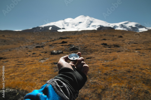 Man's hand holding a magnetic compass first-person view against the background of a high-altitude path and elbrus mountains