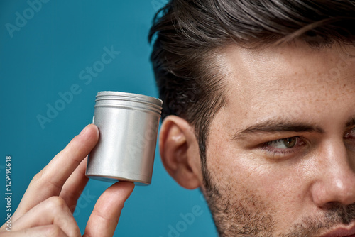 Cropped portrait of brunette guy with bristle looking away, holding silver jar with gel or cream hair styling product isolated over blue background photo