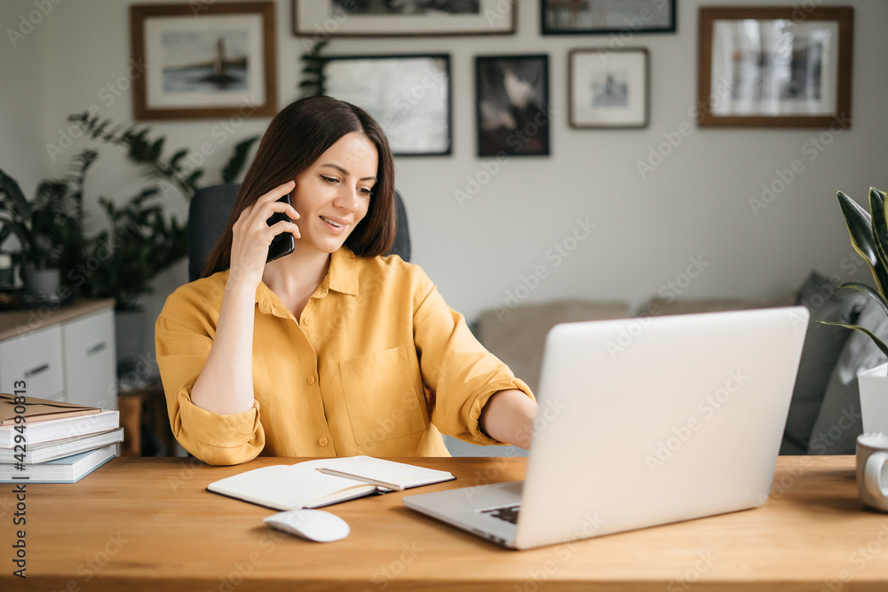Smiling business woman talking on a mobile phone and using a laptop while working at home, looking at the screen, reading a message, a young female employee advises a client, telephone conversations Smiling business woman talking on a mobile phone and using a laptop while working at home, looking at the screen, reading a message, a young female employee advises a client, telephone conversations