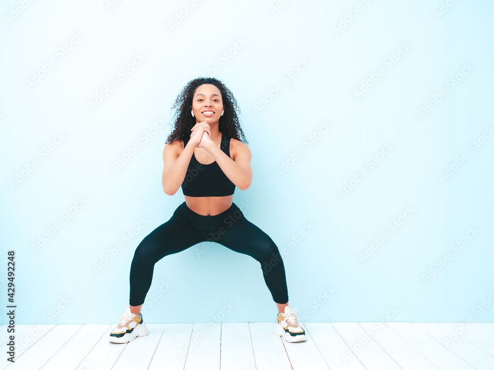 Fitness smiling black woman in sports clothing with afro curls hairstyle.She doing squats. Young beautiful model with perfect tanned body.Female in studio near light blue wall Fitness smiling black woman in sports clothing with afro curls hairstyle.She doing squats. Young beautiful model with perfect tanned body.Female in studio near light blue wall