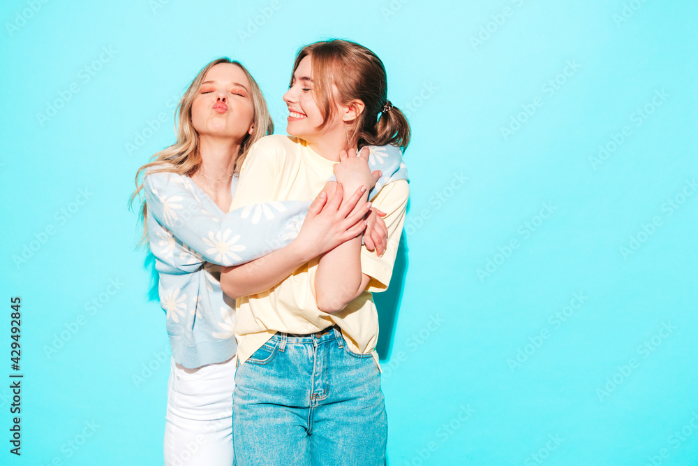 Two young beautiful blond smiling hipster female in trendy summer clothes. Sexy carefree women posing near blue wall in studio. Trendy and positive models having fun Two young beautiful blond smiling hipster female in trendy summer clothes. Sexy carefree women posing near blue wall in studio. Trendy and positive models having fun