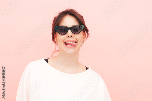 Young beautiful smiling female in trendy summer hipster clothes. Sexy carefree woman posing near pink wall in studio. Positive model having fun indoors. Cheerful and happy.Shows tongue in sunglasses