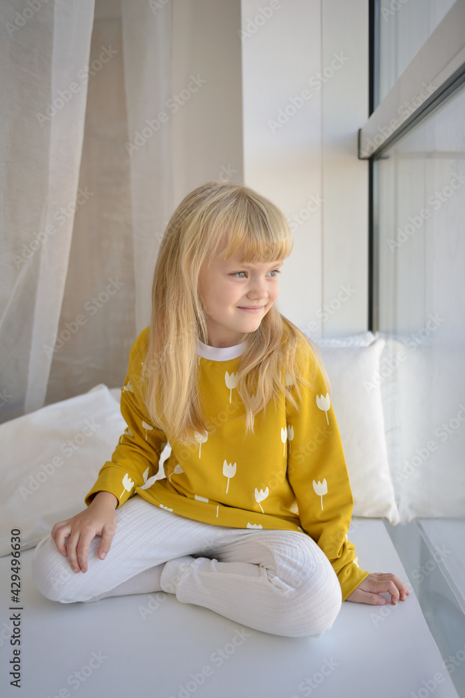 Portrait of a little girl with golden hair, blue eyes in a bright gray-yellow sports fashion suit who sits on a wide windowsill near a large window. The girl sits near the window and smiles sweetly Portrait of a little girl with golden hair, blue eyes in a bright gray-yellow sports fashion suit who sits on a wide windowsill near a large window. The girl sits near the window and smiles sweetly