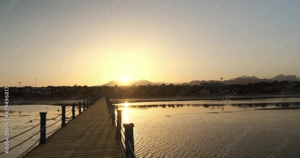 Orange sunset on wooden pier, mountain views, resort hotel, palm trees, beach. Sun sets behind mountain silhouettes, reflected in sea ripples of low tide. Vacation by sea, calm summer landscape. 4k Orange sunset on wooden pier, mountain views, resort hotel, palm trees, beach. Sun sets behind mountain silhouettes, reflected in sea ripples of low tide. Vacation by sea, calm summer landscape. 4k