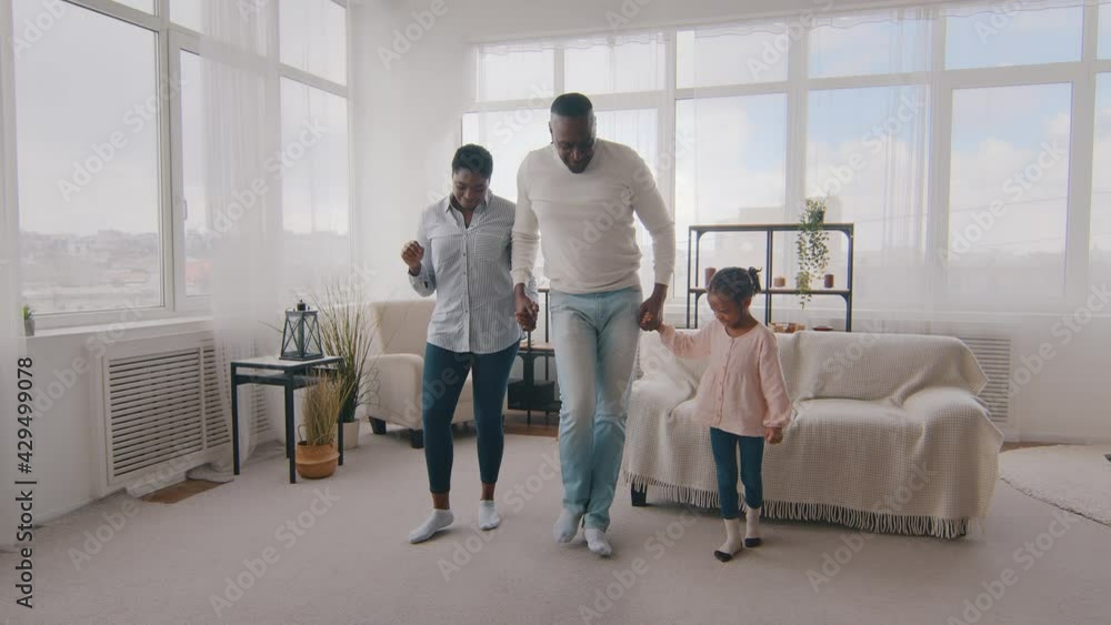 Happy afro american ethnic family adult parents with child kid dancing indoor. Black dad father teaches beloved wife mother and daughter cute girl dance shows movement with legs to music at home room Happy afro american ethnic family adult parents with child kid dancing indoor. Black dad father teaches beloved wife mother and daughter cute girl dance shows movement with legs to music at home room