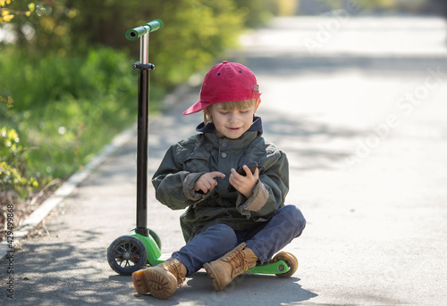a 5-year-old boy rides a three-wheeled scooter, he is tired, sitting on a scooter with a smartphone, spring background. he's wearing a parka and a cap. © Olga