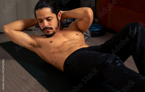 Young latin shirtless man exercising at home, doing abs.