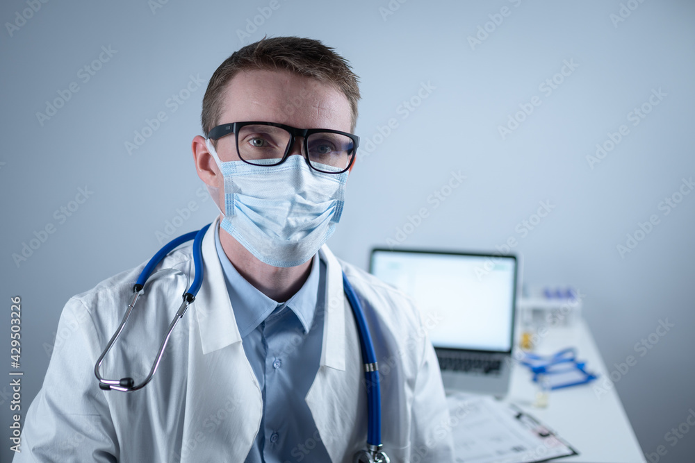 German European young doctor looking at camera wearing glasses and mask in clinic office at workplace with tired look. Topic is the hard work of medical workers during the coroavirus pandemic German European young doctor looking at camera wearing glasses and mask in clinic office at workplace with tired look. Topic is the hard work of medical workers during the coroavirus pandemic