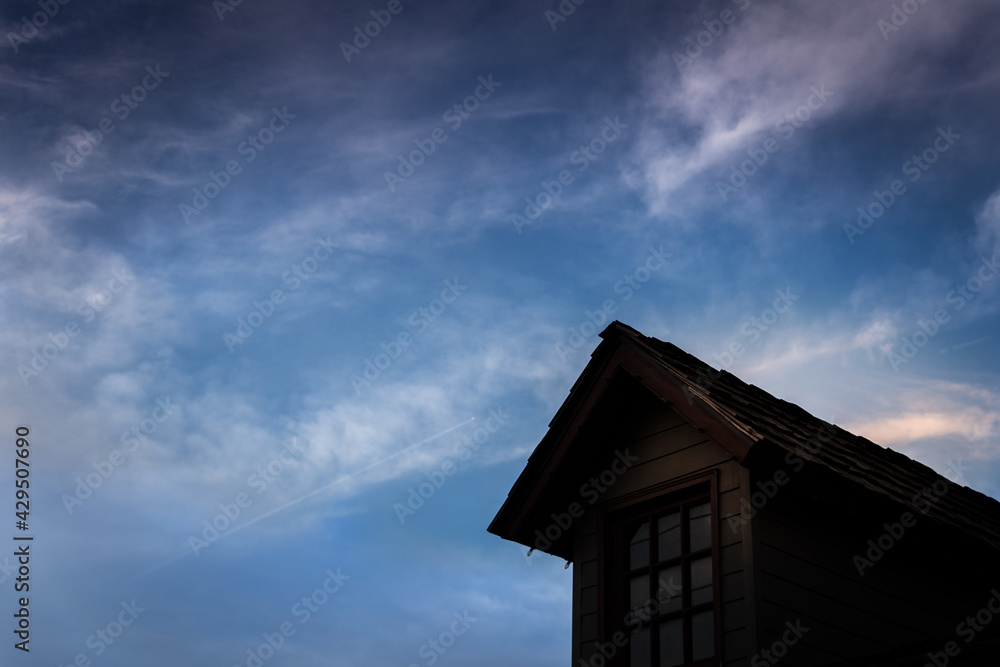 A silhouette of an old house window with it's roof under the blue sky and white clouds.