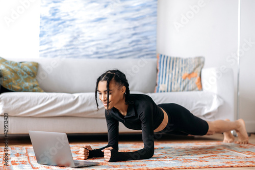 Beautiful young african american woman in sportswear, doing a plank on the floor at home, using a laptop, watching a video of fitness or yoga lessons, leading a healthy lifestyle, smiling