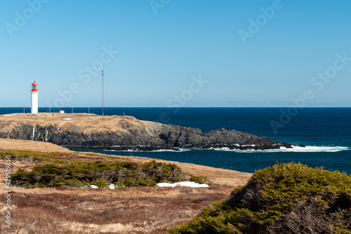 A white and red cylinder shaped concrete lighthouse with a large green glass light in the top red tower section on a cliff overlooking the blue sky and ocean with small outcrop and storage buildings.