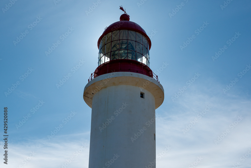 A vintage lighthouse tower with a round red metal roof.  In the center of the lighthouse is a vintage lamp made of multiple pieces of glass. On top of the white tower is a red metal wind arrow.
