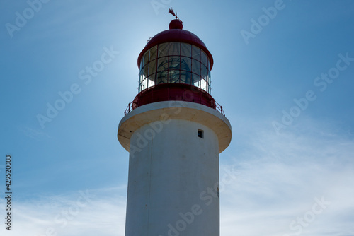 A vintage lighthouse tower with a round red metal roof. In the center of the lighthouse is a vintage lamp made of multiple pieces of glass. On top of the white tower is a red metal wind arrow.