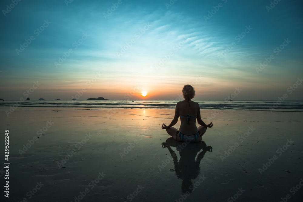 A woman sitting on beach during sunset and meditating at yoga pose.