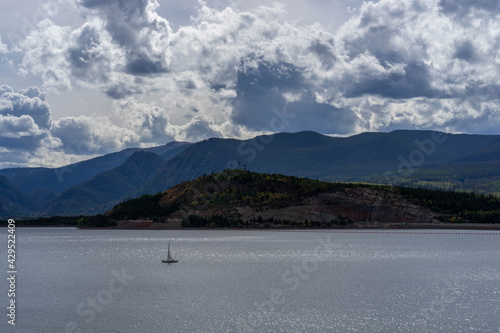Hillside and mountains with some fall foliage behind sailboat on Lake Dillon, Colorado, on partly cloudy late summer day with some sun shining through the clouds