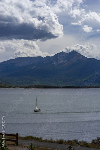 Hillside and mountains with some fall foliage behind sailboat on Lake Dillon, Colorado, on partly cloudy late summer day with some sun shining through the clouds