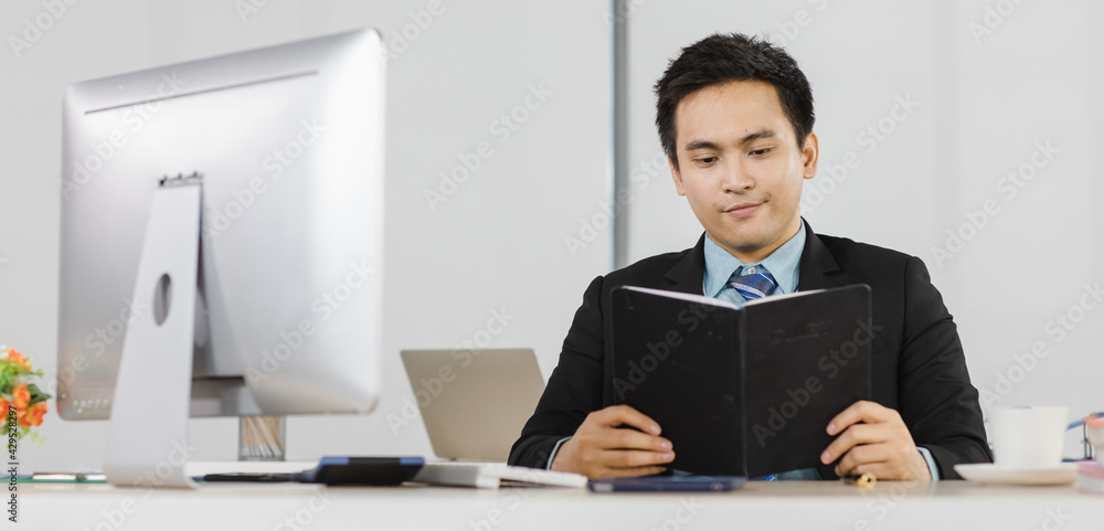 Portrait focus to adult asian businessman very smart and handsome hold and do reading note at work desk while feeling comfortable and happy smile on face. On desk have computer, calculator and tablet Portrait focus to adult asian businessman very smart and handsome hold and do reading note at work desk while feeling comfortable and happy smile on face. On desk have computer, calculator and tablet