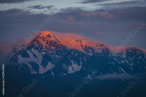 Scenic mountain landscape with great snowy mountains lit by dawn sun among low clouds. Awesome alpine scenery with high mountain pinnacle at sunset or at sunrise. Big glacier on top in orange light. © Daniil