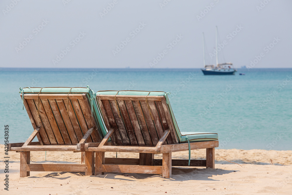 Fototapeta premium Two wooden deckchairs on a tropical sand beach overlooking the sea water and yacht. Thailand